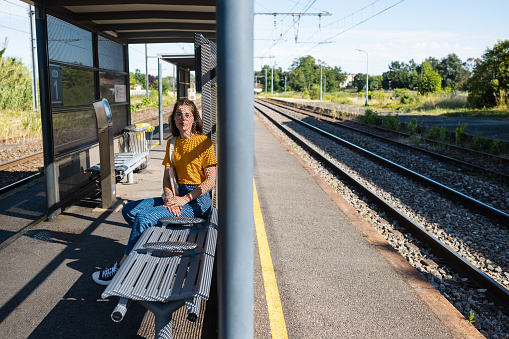 A young woman sitting on a seat on a train platform waiting for a train to arrive to take her to her next destination while on holiday in Toulouse, France. She is looking at the camera and smiling while the sun shines on her face through holes in the shelter.