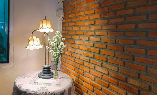 Illuminated lighting of The old vintage table lamp with daisies bouquet in glass vase on round table beside concrete and brick wall in retro restaurant