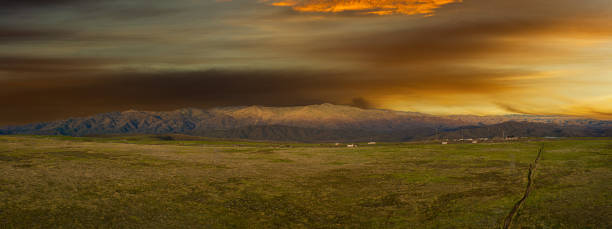 An aerial panorama of a large distant mountain under an evening sky with interstate traffic moving in front of it and a large green prairie in the foreground.