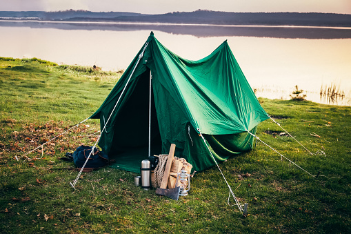 Green tent standing on the coast of the lake in the misty morning. Thermos, backpack, hatchet, firewood, ropes and kerosene lantern laying on the grass left by sleeping campers