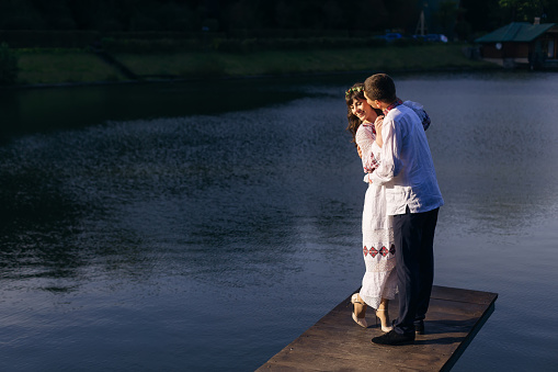 guy in an embroidered shirt hugs a girl and kiss. the girl in an embroidered dress. couple on wooden pier of lake.