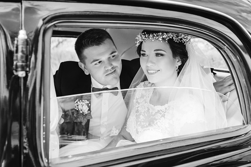 Newlyweds in festive clothes sit in a car on black and white photography