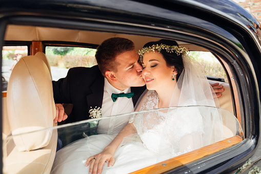 cute bride has closed her eyes and the groom kisses her on her cheek. newlyweds are sitting in the car