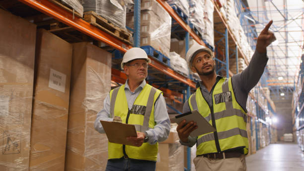 istock Employees, man and pointing at shipping cargo in warehouse for delivery or ecommerce with tablet. Logistics, boxes and factory with male person together in safety uniform for delivery with tech. 1543425328