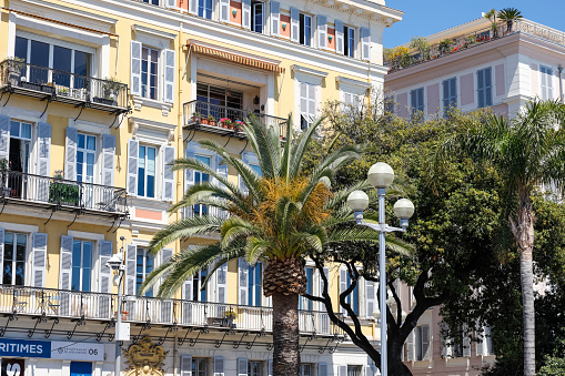 Nice, France - May 3, 2023: Mediterranean architecture on the example of a building facade with balconies and shutters in a city where palm trees and other trees and plants grow next to the buildings