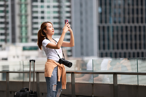 Young Asian Photographer using a smartphone and holding the digital camera at the city in high building background.