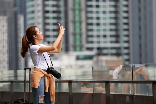 Young Asian Photographer using a smartphone and holding the digital camera at the city in high building background.