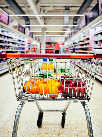 Selective focus color image depicting a shopping cart filled with fresh groceries and fruit in the supermarket aisle. Focus on the shopping cart in the foreground with the aisle defocused beyond.
