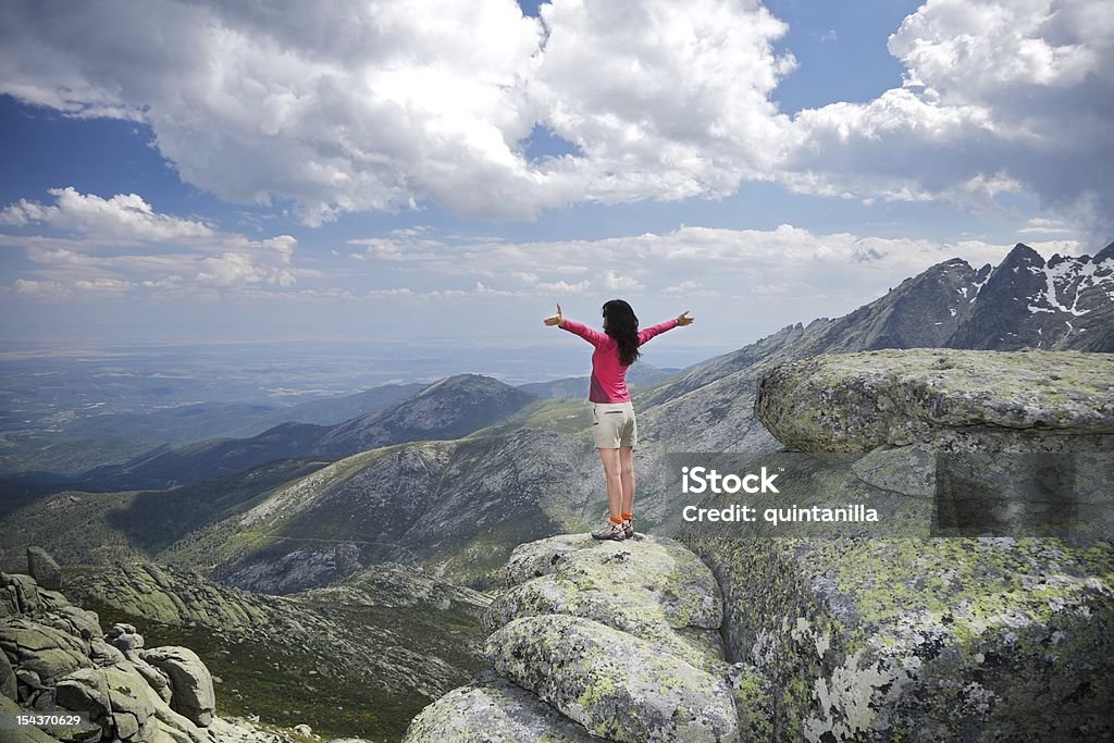 open arms cross woman at top summit mountains touch clouds Backwards side brunette woman red cardigan brown khaki shorts open arms like fly. She is a trekking hiking sport people in summer spring time. She is on the top of a grey and green moss rock in summit of mountains in Gredos area Avila, Castile, Spain Europe One Woman Only Stock Photo