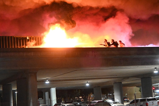 Oakland, CA USA - Dec 3, 2022: Firefighters battle a deadly truck fire on an elevated freeway in Oakland, CA.\n\nAfter being struck by a stolen car, this truck overturned, hit the railing and burst into flames. The driver was unable to escape and died on scene. The stolen car was abandoned and the occupants picked up by another car that drove off.
