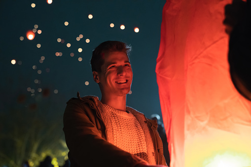 Confident smiling man lighting up a Chinese lantern at event at night. Happy man experiencing and enjoying different culture tradition.