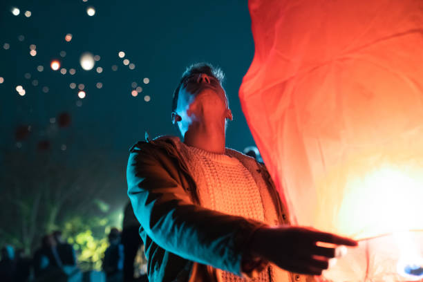 Young smiling man holding a paper sky lantern at Chinese event at night while looking at the illuminated sky