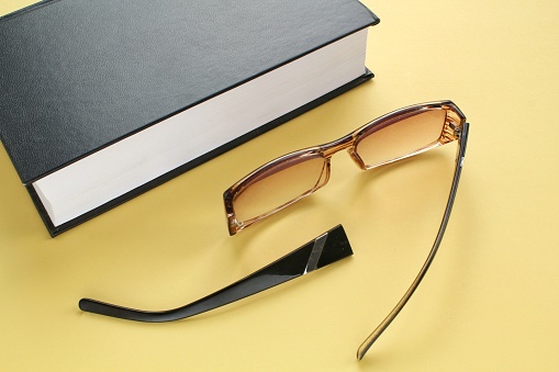 Old broken eyeglasses with damaged legs and books on yellow background. Poor eyesight. Repair concept. Idea of health. Failure optic eyewear. Breakage of vision correction glasses. Close up, flat lay