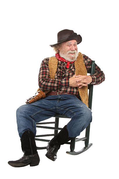 Friendly old cowboy sits in rocking chair Classic old west style smiling cowboy with felt hat, grey whiskers, revolver. He sits in a rocking chair. Isolated on white, vertical, copy space. checkered cowboy boots stock pictures, royalty-free photos & images