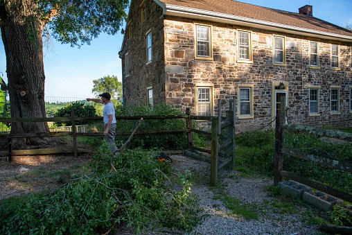 Home owner stands by an old stone home with a wooden split rail fence, inspecting broken downed branches in the yard from a storm. He surveys the damage by a tall tree..