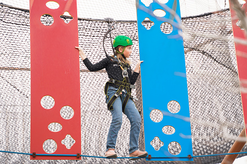 Teenage teen girl in climbing harness equipment, green sports safety helmet. Rope amusement park. Fastening attaching carabiner to safety rope. Hanging obstacle course. Kids children's attraction.