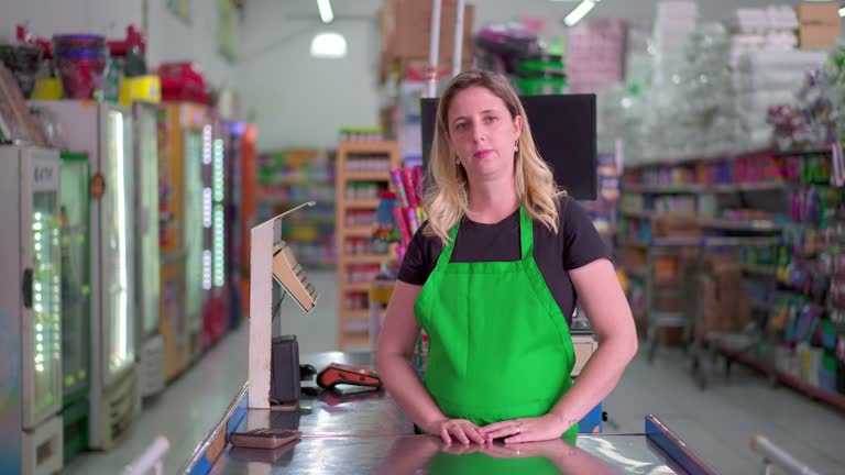 Female employee of Supermarket standing by cashier checkout looking at camera with neutral expression wearing green apron. Portrait of staff of Grocery Store