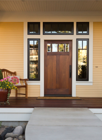 Front view of a wooden front door on a yellow house with reflections in the window and a wide view of the porch and front walkway. Vertical shot.