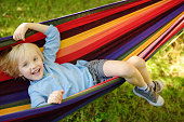 Cute little blond caucasian boy relaxing and having fun in multicolored hammock in backyard or outdoor playground. Summer active leisure for kids. Child swinging on hammock.