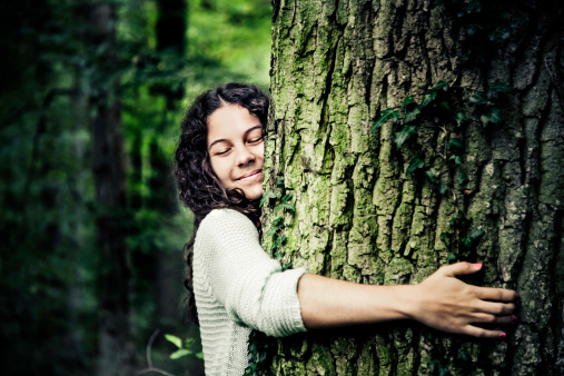 Pretty natural girl with black curled hair leaning on a tree trunk in a beautiful lush green forest