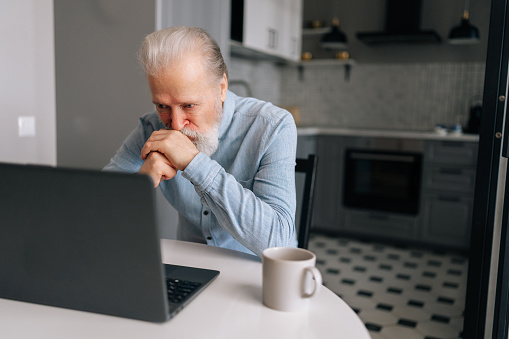 Portrait of pensive bearded senior older businessman thinking about job and looking on screen of laptop sitting at table with coffee cup. Worried aged gray-haired man put chin on hands feels abandoned
