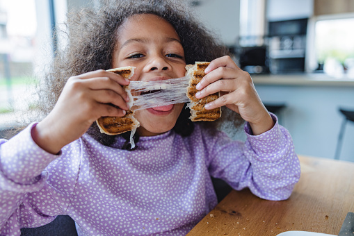 A portrait of a cute young black girl in purple pajamas having fun while breaking her toast sandwich in half. She is smiling while playing with her food. The young girl is sitting on a chair next to a wooden dining table in a beautiful modern kitchen with big windows.