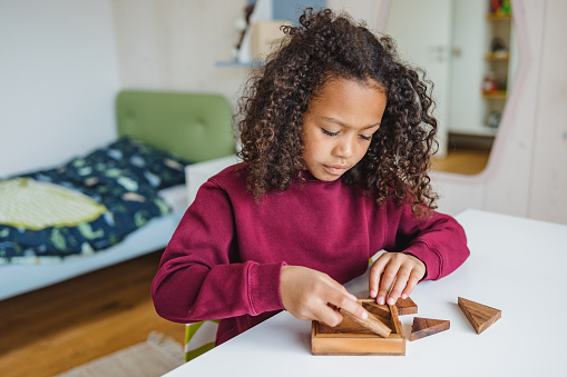 A portrait of an adorable little black girl playing in her room and sitting by a white desk. She is playing with a puzzle toy and looks focused on solving the puzzle. She has beautiful long curls and is wearing a burgundy sweatshirt. The kid's room has modern furniture that is white blue and green.