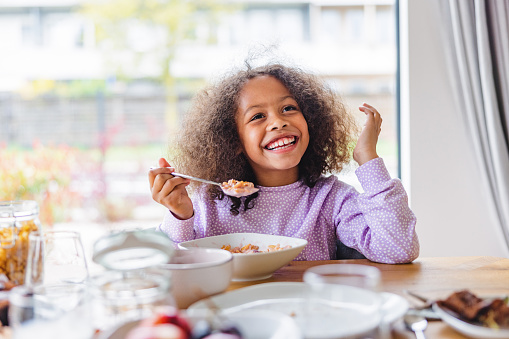 Happy cute young black girl smiling while eating a bowl of cereal. She is sitting on a chair next to the wooden table that is full of plates and food. The young girl has cute curly hair and is wearing a purple pajama. In the back there is a big window that allows natural light into the modern apartment.