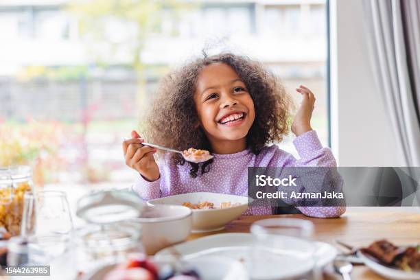 Adorable Black Girl With Curly Hair Smiling While Eating Breakfast In A Bright Kitchen Stock Photo - Download Image Now