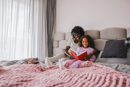 A happy black mom and her young daughter enjoying spending time together while lying in a big double bed and reading a red book. They are both dressed in pajamas. The mother has her hand around her daughter. They are both happy and smiling. The bed that they are lying in is located next to a big window in a modern bedroom.