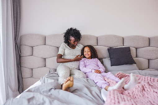 Black pregnant mother looking at her young little girl while they are both cuddling in a big cosy bed in a modern bedroom. The mother has her hand around her young daughter. They are lying and cuddling in a bed full of pillows and blankets. The girls are wearing their pajamas and both have long curly hair.