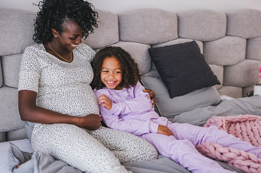 Happy black pregnant mother hugging her young daughter while they are lying in a comfortable bed. They are both wearing cute pajamas and have long curly hair. The mother is looking at her smiling daughter while having her arm around her. They are surrounded by blankets and pillow.