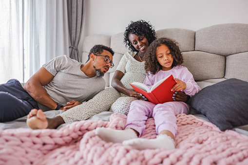 Young black girl reading a book to her happy black parents. The black family is lying together on a double bed and cuddling while the daughter is trying to learn how to read. The daughter has a red book in her hands. The mother is smiling while looking at the child. They are all wearing pajamas. They are located in luxurious bedroom with modern interior and big windows.