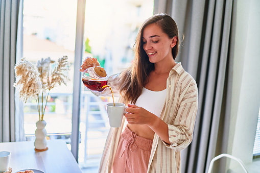 Happy young cute joyful smiling satisfied woman pouring hot tea into cup from glass teapot at home