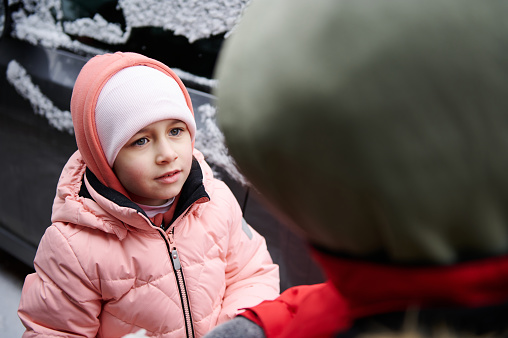 Portrait of a Caucasian beautiful preschool child girl in warm winter clothes, talking to someone older standing near a gray snow covered car. Childhood. Children. Innocence. Winter time. Confidence