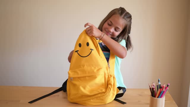 Beautiful Smiling Girl Packing The School Supplies In The Yellow Backpack.