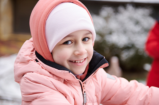 Close-up Caucasian beautiful little child girl 6 years old, dressed in pink warm winter clothes, smiling cutely, looking at camera, standing in the snow covered backyard. Happy kids. Childhood concept