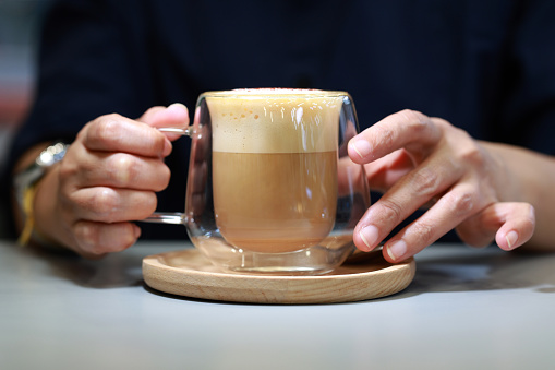 In a close-up shot, a woman's hands delicately hold a cup of latte coffee served in a double-layer glass cup at a café.