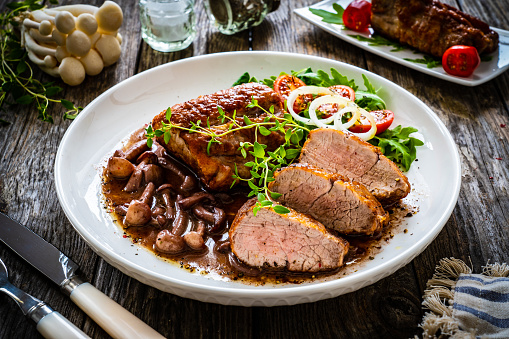 Roast pork loin, mushrooms in sauce and fresh vegetables on wooden table