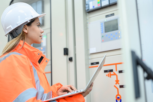 Asian electrical technician worker woman with personal safety equipment using laptop computer operating or check status from control panel switch board in control room of high voltage electric transformer station. Industrial business occupation worker concept.