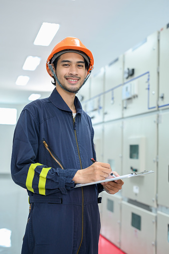 Portrait of Asian electrical technician worker smiling and looking at camera while working in control room of high voltage electric transformer station. Industrial business occupation worker concept.