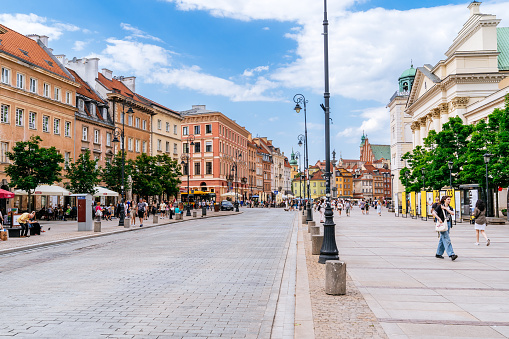 A view of Krakowskie Przedmieście street in Warsaw's Old Town Center (Stare Miasto w Warszawie, Warszawska Starówka). In the frame, two notable landmarks are visible. On the right side is Kościół Akademicki św. Anny (St. Anne's Academic Church), a historic religious building with distinctive architectural features. On the background is Plac Zamkowy (Castle Square), an open public space with a notable historical significance. The street itself is bustling with pedestrians and features a mix of architectural styles. This photograph offers a glimpse into the architectural and cultural richness of Krakowskie Przedmieście street, showcasing the presence of Kościół Akademicki św. Anny and Plac Zamkowy within the surrounding urban landscape.