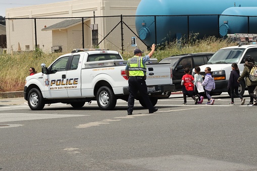 South San Francisco, United States – May 25, 2023: A uniformed police officer gesturing to the pedestrian kids to cross the street