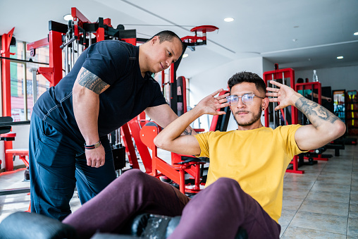 Young man doing sit-ups with help of personal trainer at gym