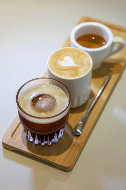 At a coffee testing event, a skillful barista places a wooden tray on the counter, artfully serving three different types of coffee. The tray holds three cups, each containing a unique coffee variant - espresso, latte, and ice black coffee.