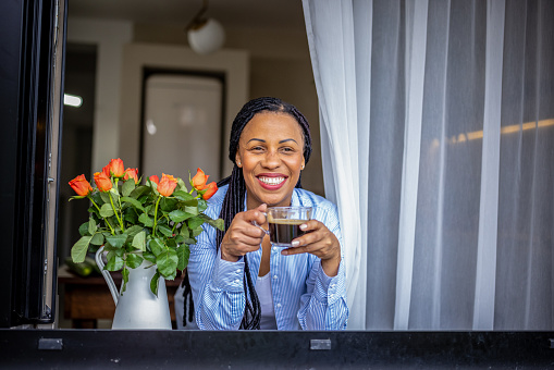 Photo of American woman drinking a cup of coffee while standing by the the window at home.