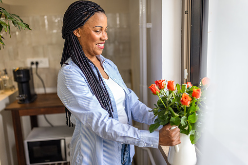 Photo of smiling afro-american woman putting vase with fresh roses on the window at home.