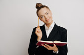 Smart female worker generates business ideas with red notebook, pencil, and smile against light wall.