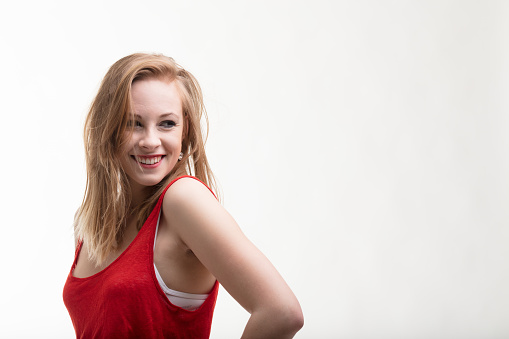 Young woman with fair skin and strawberry-blonde hair wearing a vibrant red dress. She smiles, then laughs, accustomed to meeting others' gazes. Half-length portrait on a white background with ample c