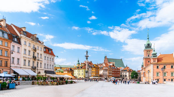 Panoramic View Of Warsaw Old Town Center - Castle Square And Zamek Królewski w Warszawie (Royal Castle in Warsaw) Against Blue Sky. A panoramic view of Castle Square (Plac Zamkowy) in Warsaw, Poland. The image captures the various landmarks, buildings, and restaurants that make up the square. Notably, on the right side of the photo, Zamek Królewski w Warszawie (Royal Castle in Warsaw) is clearly visible. The facades of the buildings exhibit the architectural style of the area. People can be seen leisurely strolling along the square, adding a sense of activity to the scene. The sky above is adorned with a vibrant blue hue, with scattered clouds indicating a pleasant summer day. This photo provides an accurate representation of the significant landmarks, diverse buildings, bustling restaurants, and the presence of Zamek Królewski w Warszawie in Castle Square, Warsaw, Poland. warszawa stock pictures, royalty-free photos & images